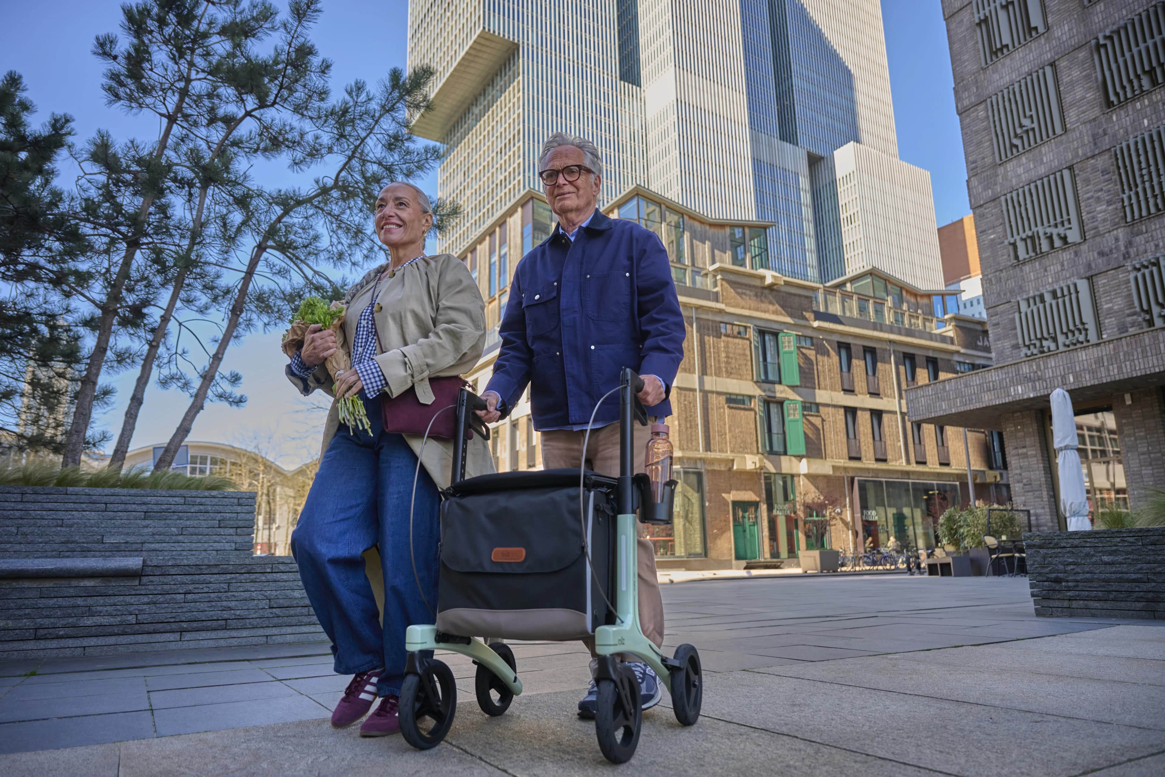 Een vrouw en een man wandelen met boodschappen en een Rollz Air rollator door de stad.
