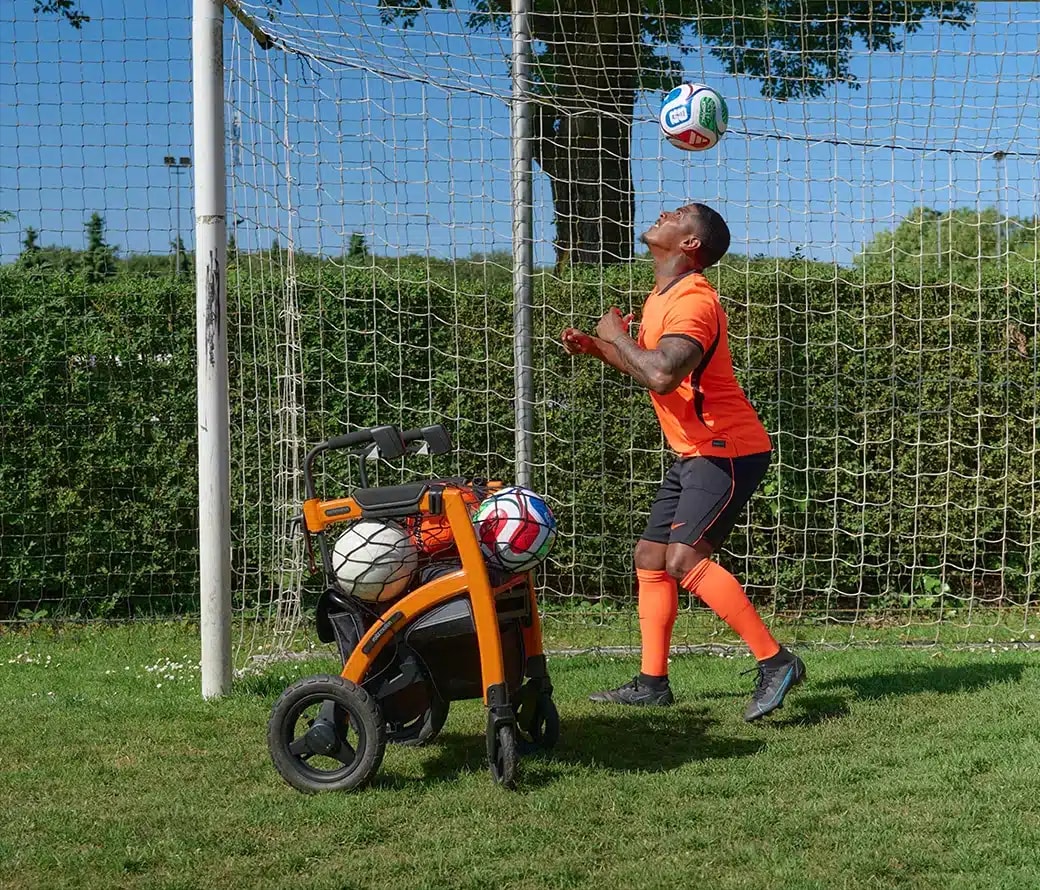 Een voetballer in een oranje tenue kopt een bal voor het doel, met de oranje Rollz Motion Performance vol ballen naast zich.