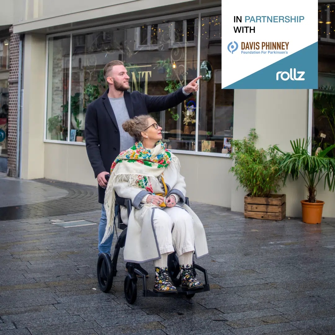 Een man duwt een vrouw in een Rollz Motion door een winkelstraat, met het logo In partnership with Davis Phinney.