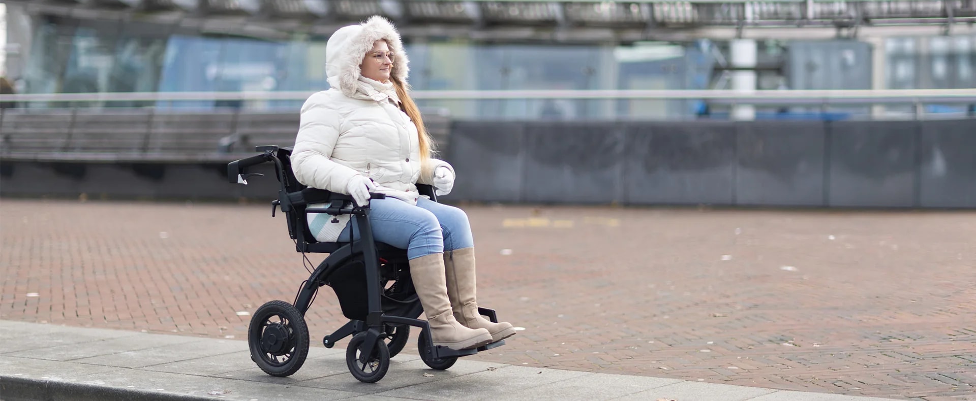 Een vrouw in een witte winterjas met bontkraag zit in de Rollz Motion Electric op een boulevard.