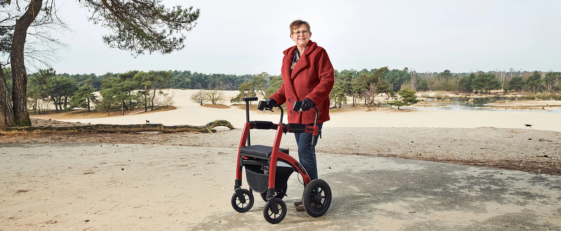 Een vrouw in een rode wollen jas staat met de rode Rollz Motion Performance op een zandvlakte aan de rand van het bos.