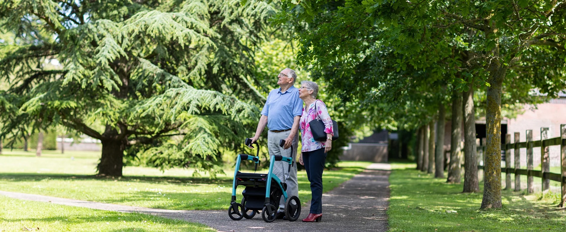 Een ouder echtpaar staat met de turquoise Rollz Motion op een parkpad onder hoge bomen en kijkt omhoog.