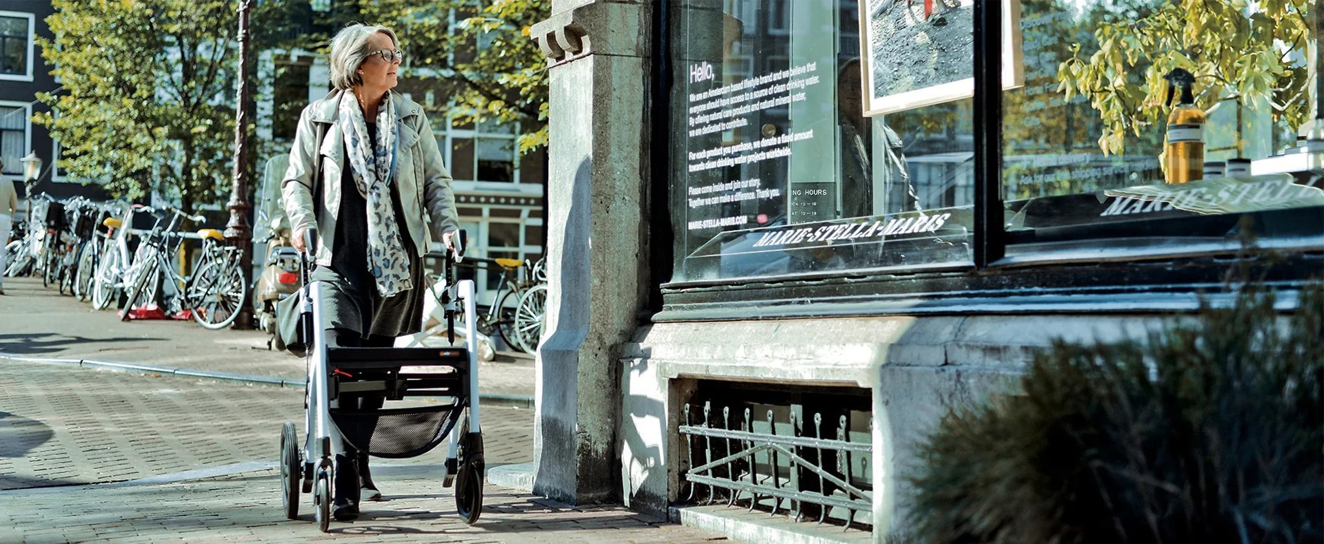 Een vrouw met grijs haar loopt met een witte Rollz Motion langs een etalage aan een Amsterdamse gracht, met geparkeerde fietsen op de achtergrond.