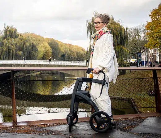 Een vrouw staat met de Rollz Motion Rhythm Parkinson rollator bij een brug over het water