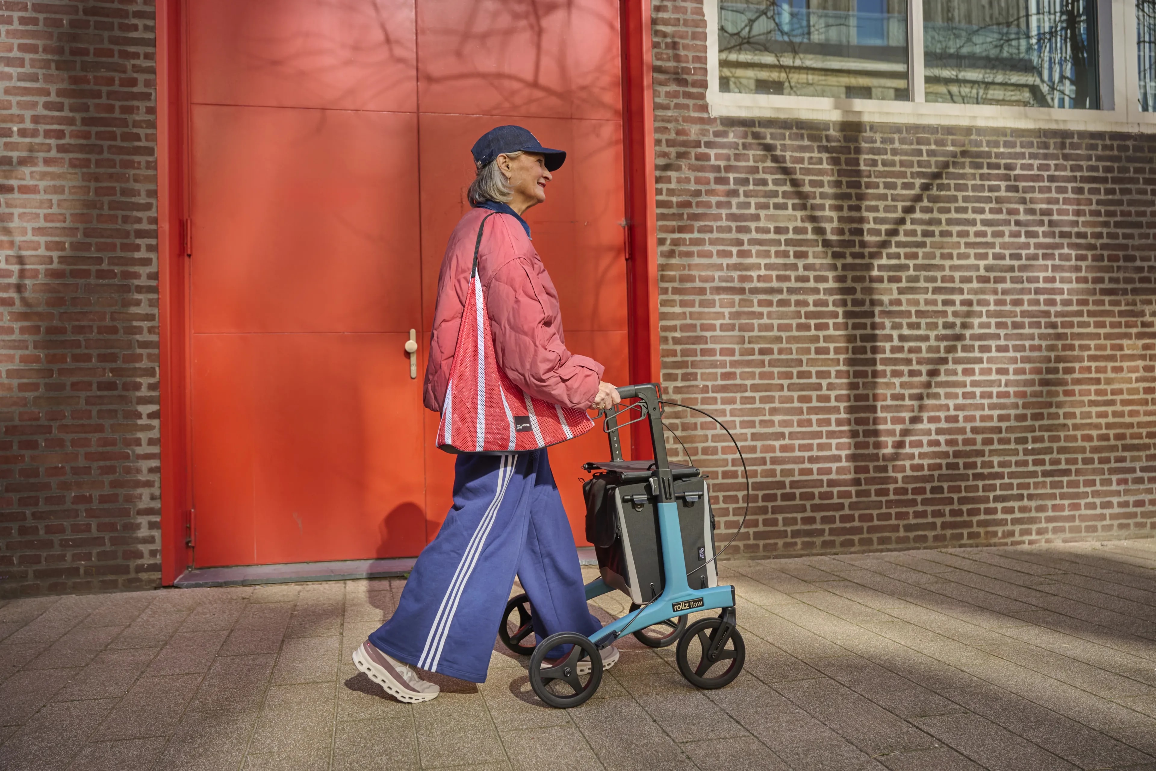 Een vrouw met pet en rode tas loopt met een blauwe Rollz Flow rollator langs een rode deur in de stad.