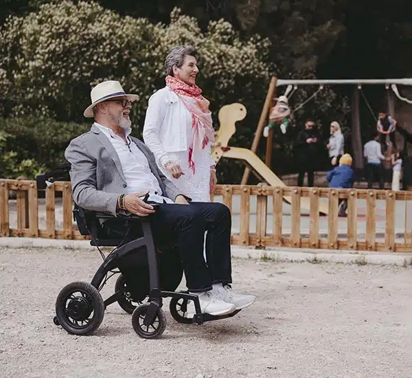 Een man met een hoed zit in de Rollz Motion Electric bij een speeltuin, een vrouw staat lachend naast hem.