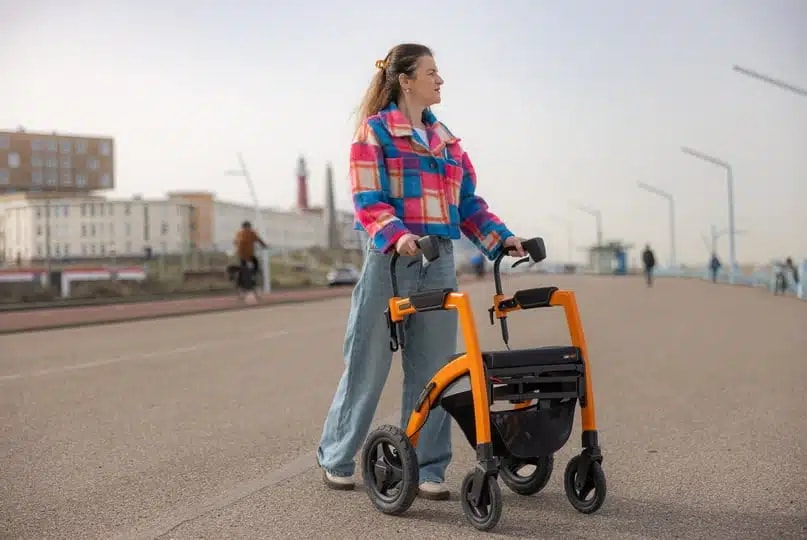 Een jonge vrouw in een geruit jack loopt met de oranje Rollz Motion Performance over een boulevard aan zee.