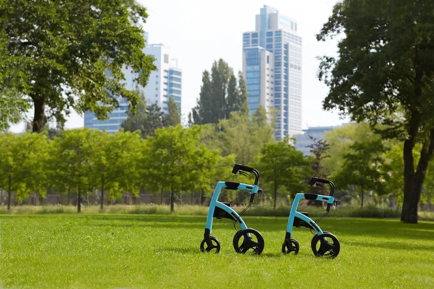 Twee lichtblauwe Rollz Motion rollators staan op een grasveld in het park, met hoogbouw op de achtergrond.