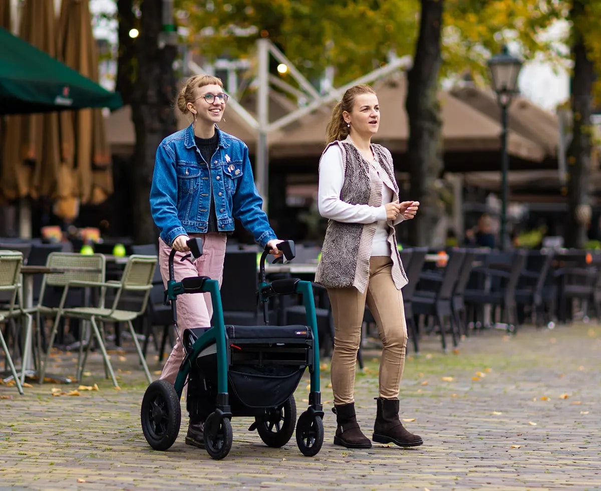 Twee vrouwen wandelen over de kinderkopjes van een plein met terrassen, een van hen met de groene Rollz Motion Performance.