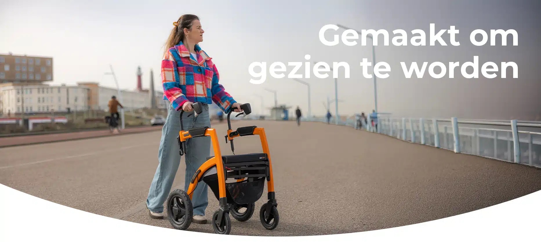 Campagnebeeld van de Rollz Motion Performance Dutch Orange: een vrouw in een geruite jas met de oranje rollator op een boulevard aan zee, naast de tekst “Gemaakt om gezien te worden”.